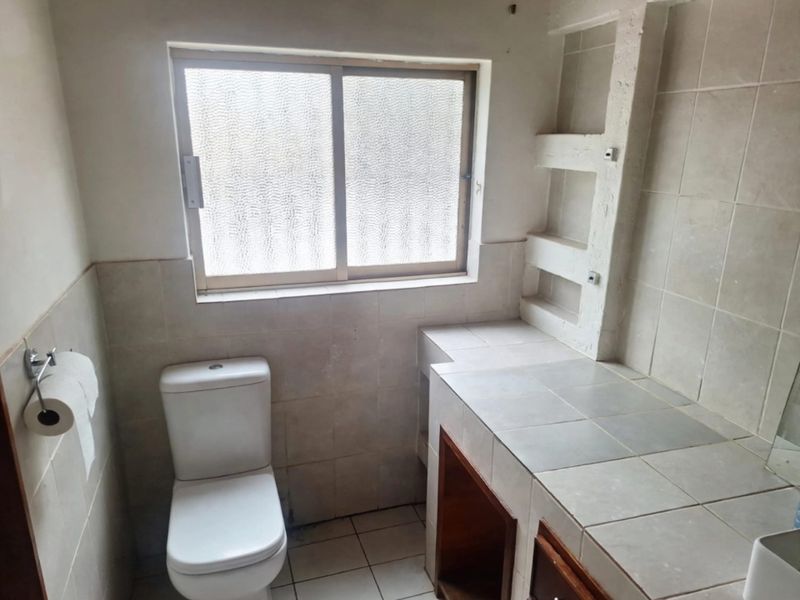 A functional bathroom featuring a toilet, tiled vanity with built-in shelving, and a sliding window, requiring cosmetic updates to improve its overall condition.
