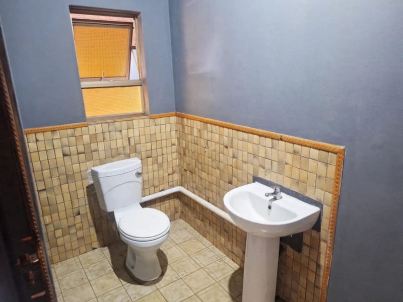 A compact bathroom featuring a pedestal basin, toilet, and tiled finishes. The space requires modernization, particularly regarding the exposed surface-mounted plumbing.