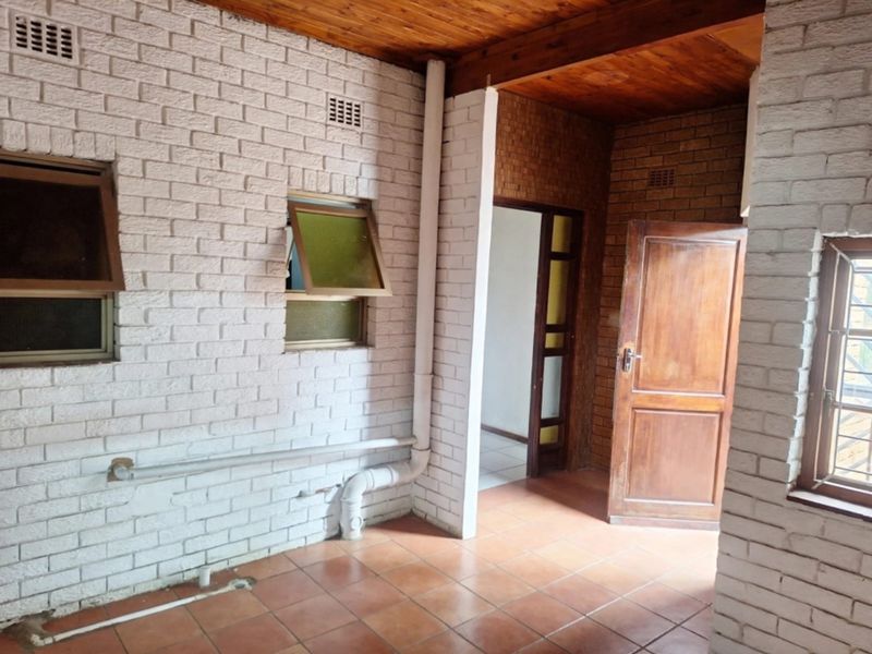 A functional laundry area featuring painted brick walls, tiled flooring, and a timber ceiling, with exposed plumbing infrastructure and direct access to the interior.