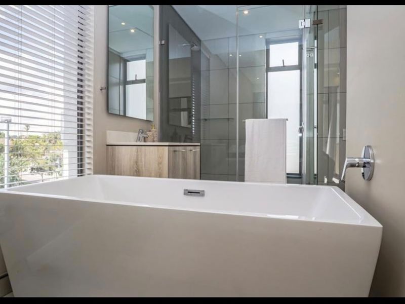 Modern bathroom featuring a sleek freestanding bathtub, glass-enclosed shower, and contemporary vanity with neutral tiling and large windows.