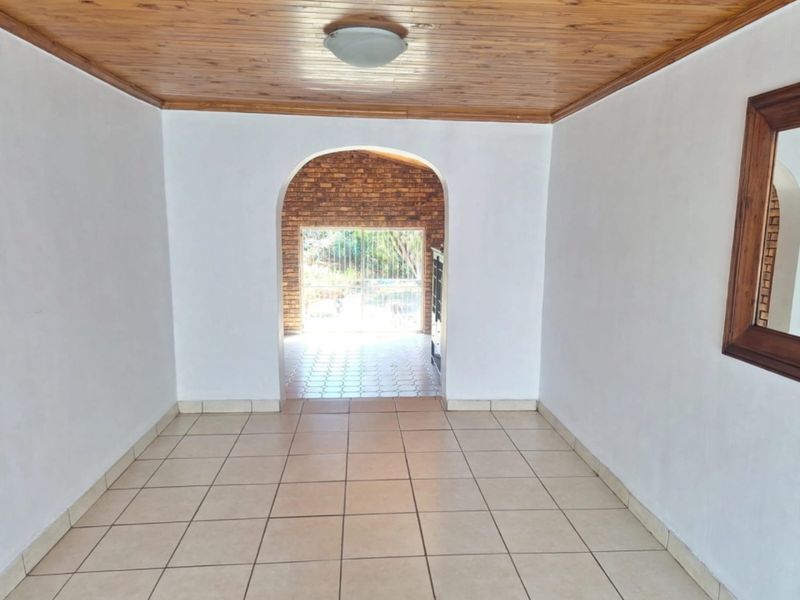 An empty living area featuring tiled floors and a timber ceiling, leading through an archway to an outdoor patio space.
