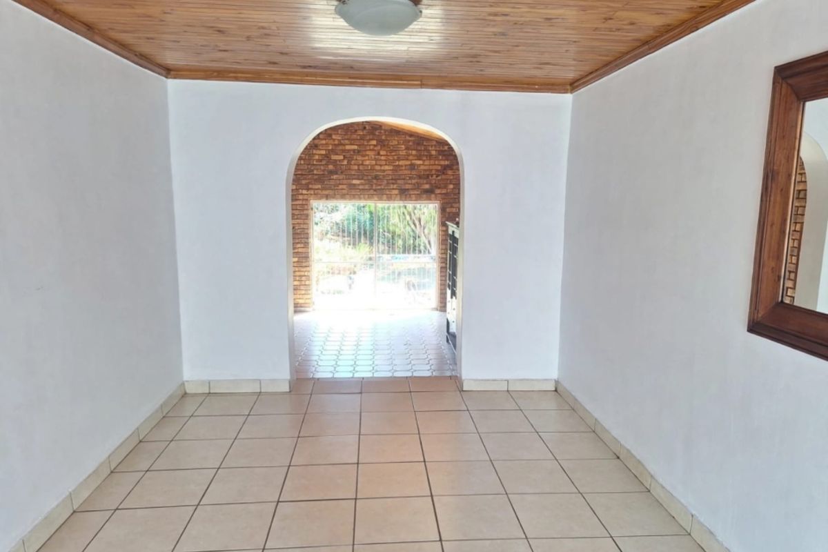 An empty living area featuring tiled floors and a timber ceiling, leading through an archway to an outdoor patio space.