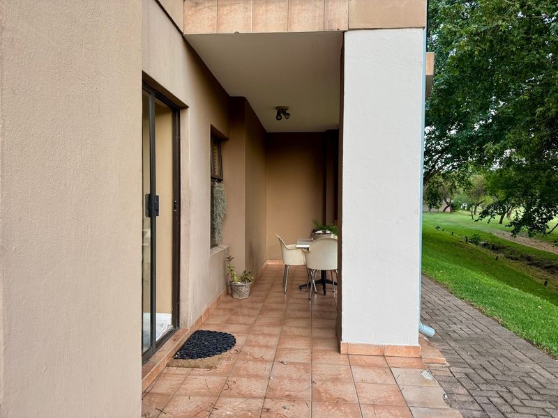 A covered patio area featuring tiled flooring and a sliding glass door entrance, overlooking a green garden space.
