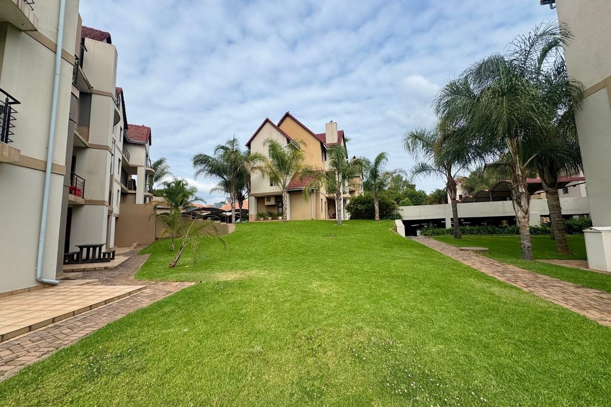 A well-maintained communal garden area between apartment blocks, featuring paved walkways, manicured lawns, and established palm trees.