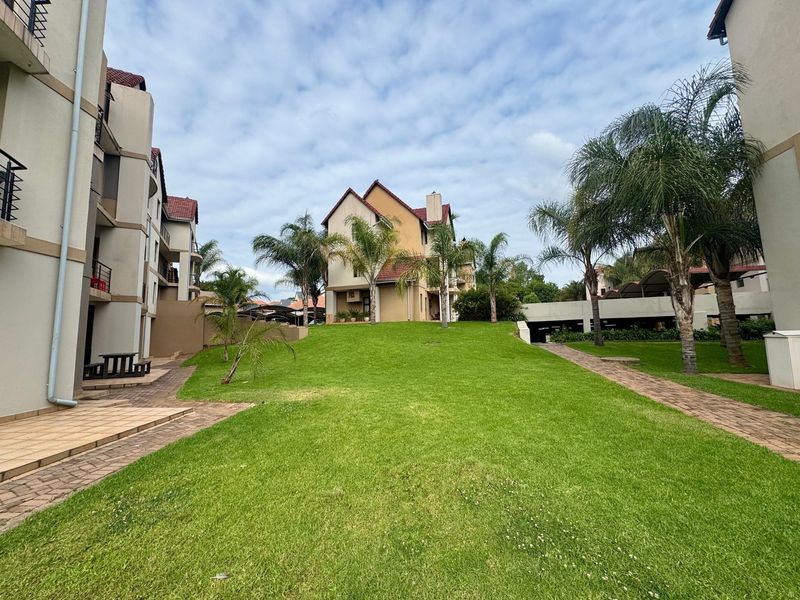 A well-maintained communal garden area between apartment blocks, featuring paved walkways, manicured lawns, and established palm trees.