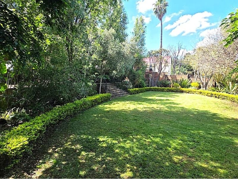 A well-maintained, lush green garden featuring mature trees, manicured hedging, and stone steps leading to an elevated area.