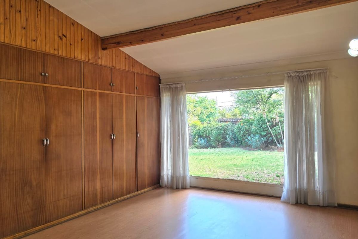 Spacious bedroom featuring extensive built-in wooden cabinetry, exposed timber ceiling beams, and a large window overlooking the garden.
