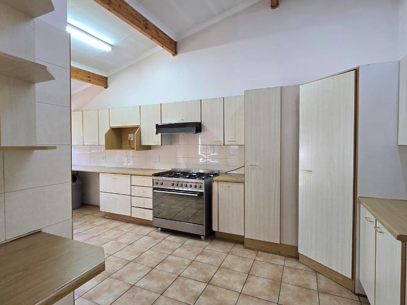 A spacious, empty kitchen featuring light-toned built-in cabinetry, a gas stove with extractor fan, tiled flooring, and high ceilings with exposed timber beams.