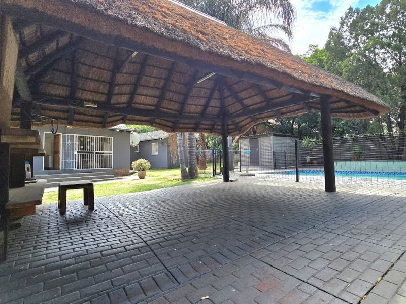 Covered outdoor patio featuring a traditional thatched roof, paved flooring, and a built-in braai, overlooking the swimming pool and garden area.