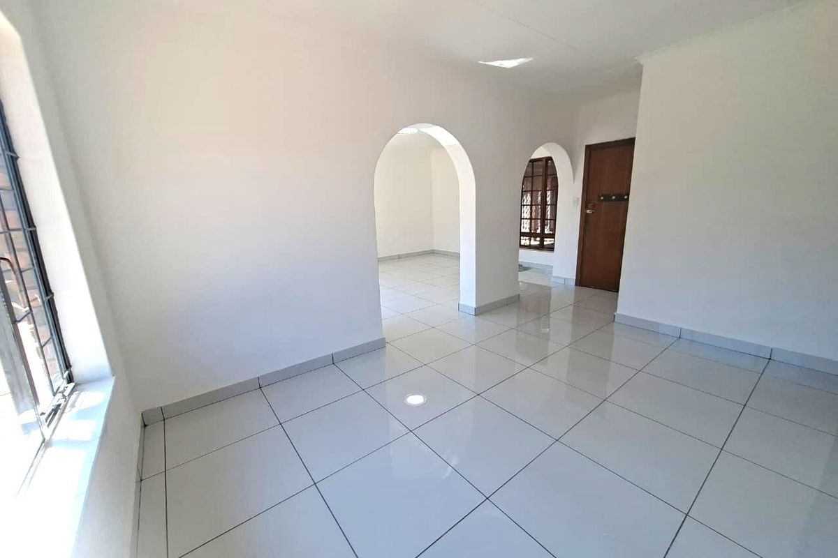 A bright, empty living area featuring white tiled floors, arched architectural transitions, and large windows with security bars.