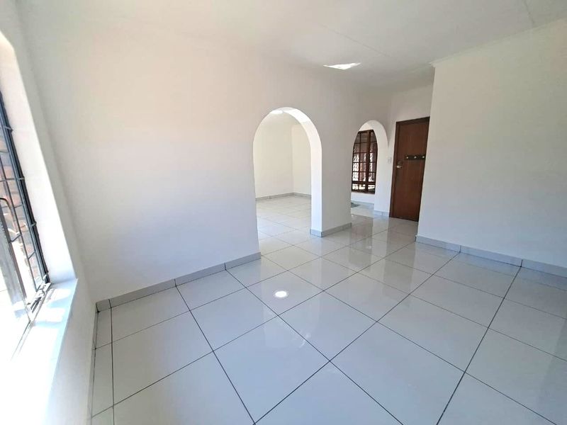 A bright, empty living area featuring white tiled floors, arched architectural transitions, and large windows with security bars.