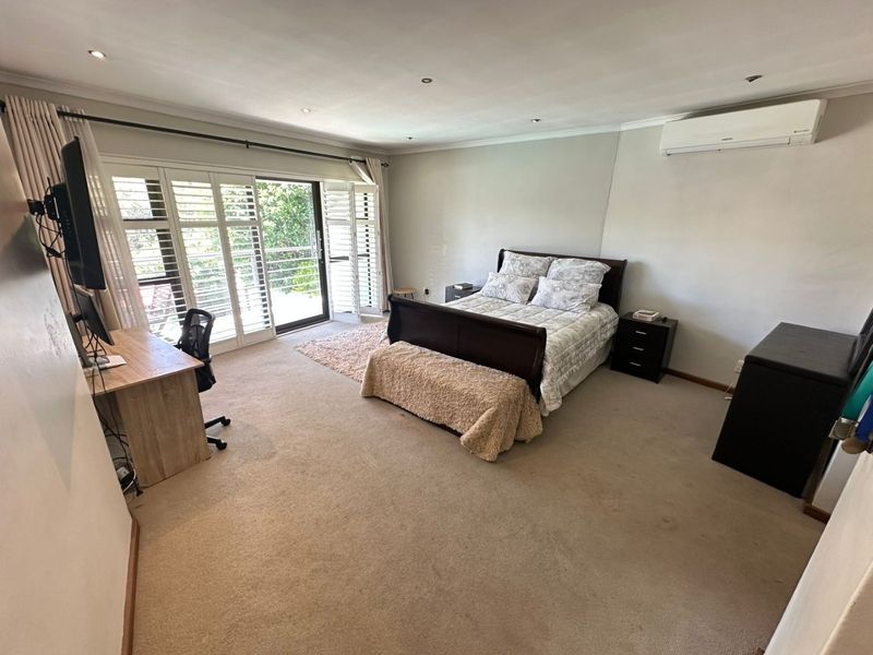 Spacious bedroom featuring carpet flooring, recessed downlights, a wall-mounted air conditioner, and plantation shutters opening to an outdoor area.