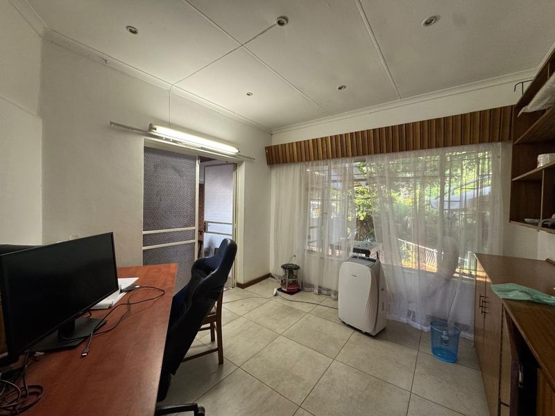 A functional office space featuring tiled flooring, built-in shelving, and large windows with security bars, offering potential for a modern workspace update.