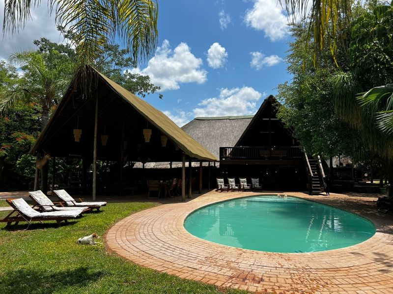A spacious outdoor pool area featuring a large swimming pool, brick paving, and a covered thatched patio structure with an upper-level wooden balcony.