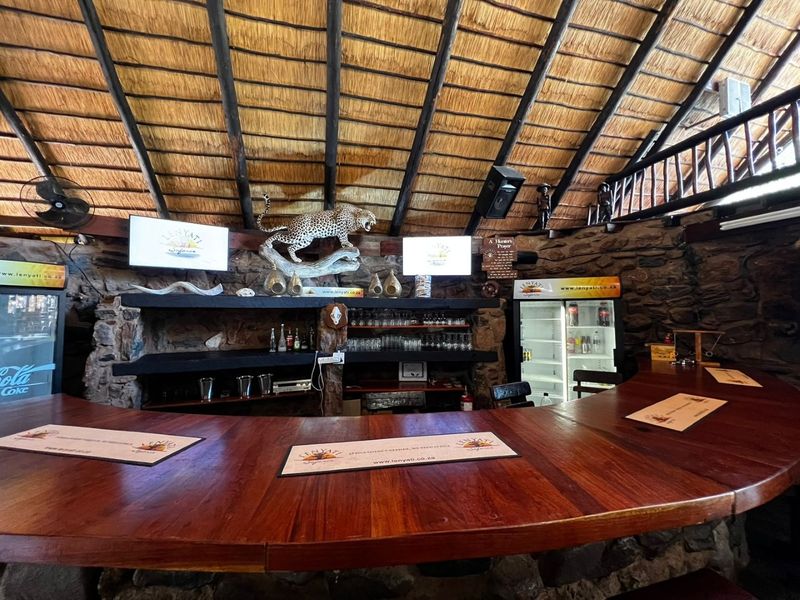 A rustic bar area featuring a curved wooden counter, natural stone walls, and a traditional thatched roof with exposed timber rafters.