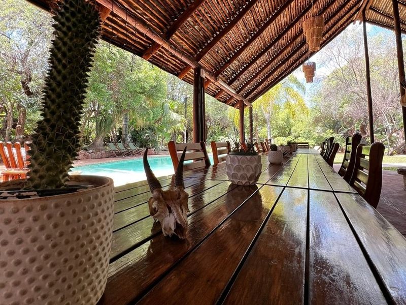A spacious covered outdoor patio featuring a rustic thatched roof and timber structure, overlooking a private swimming pool and lush garden setting.