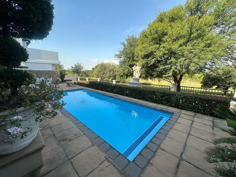 A well-maintained rectangular swimming pool set within a paved patio area, featuring landscaped garden borders and metal perimeter fencing.
