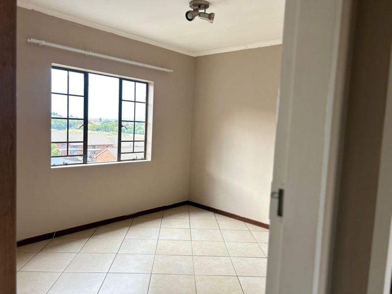 A bright, empty bedroom featuring neutral-toned walls, tiled flooring, and a steel-framed window with a view of the surrounding neighborhood.