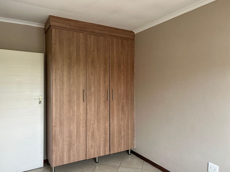 A clean bedroom interior featuring a modern wood-finish built-in wardrobe, neutral wall tones, and tiled flooring.