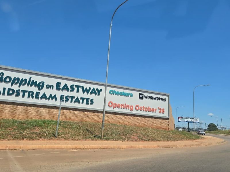 A street-level view of a brick boundary wall featuring a large commercial billboard advertising a new shopping center development in Midstream Estates.