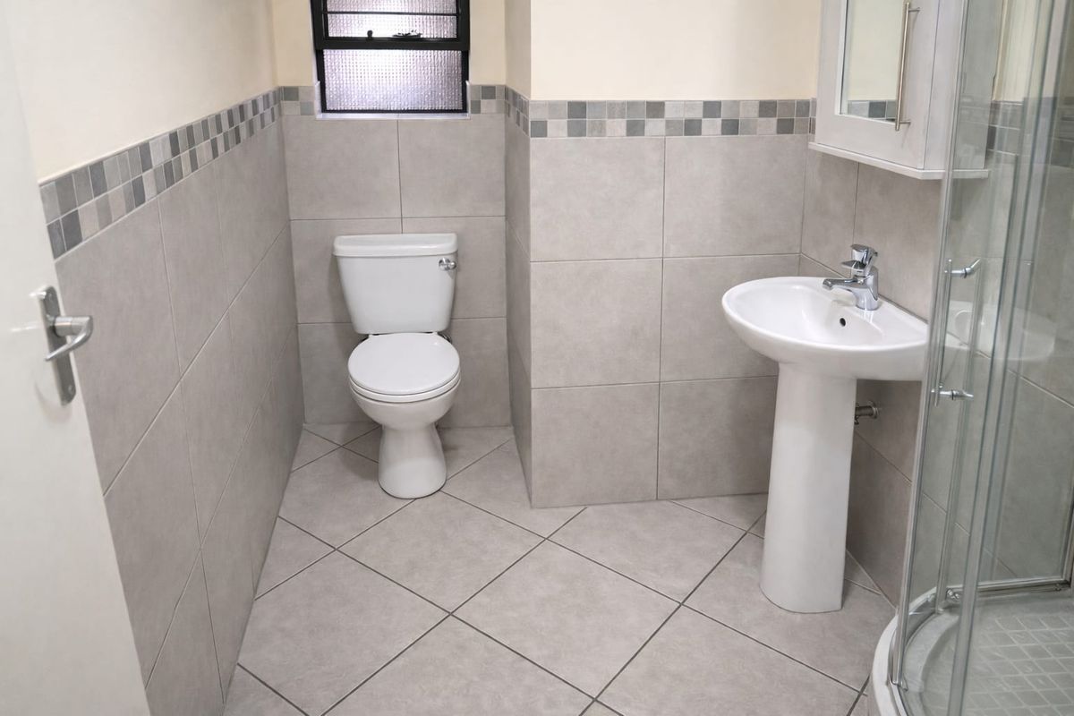A clean, modern bathroom featuring a pedestal basin, toilet, and a glass-enclosed shower, finished with neutral wall and floor tiling.