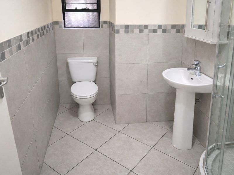 A clean, modern bathroom featuring a pedestal basin, toilet, and a glass-enclosed shower, finished with neutral wall and floor tiling.