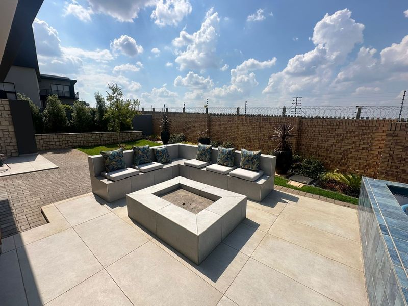A modern outdoor patio featuring a built-in fire pit and integrated seating area, set within a landscaped garden with a brick boundary wall.