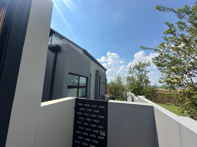 A modern rear exterior view featuring clean plastered walls, dark aluminium window frames, a secure metal gate, and an integrated air conditioning unit.
