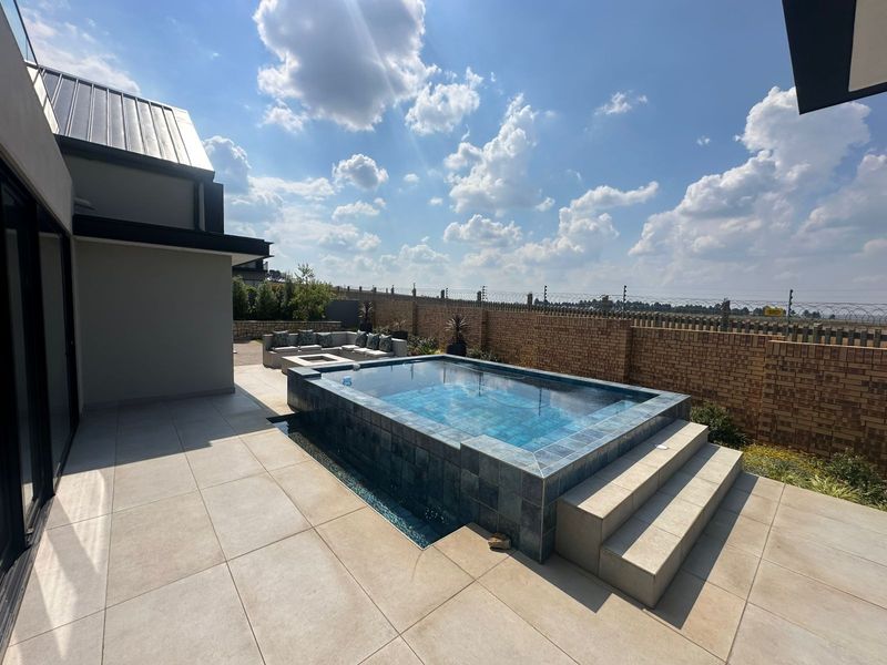 A modern outdoor pool area featuring a raised tiled swimming pool, expansive patio space, and secure brick boundary wall with electric fencing.