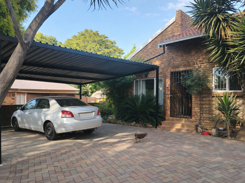 Front exterior view of a brick house featuring a paved driveway, a modern metal carport, and secure entrance gate.