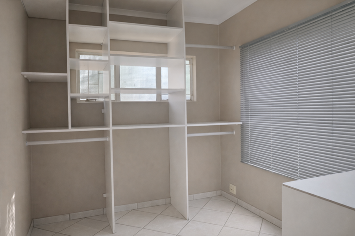 A functional bedroom space featuring extensive built-in white shelving and hanging rails, tiled flooring, and venetian blinds for privacy.