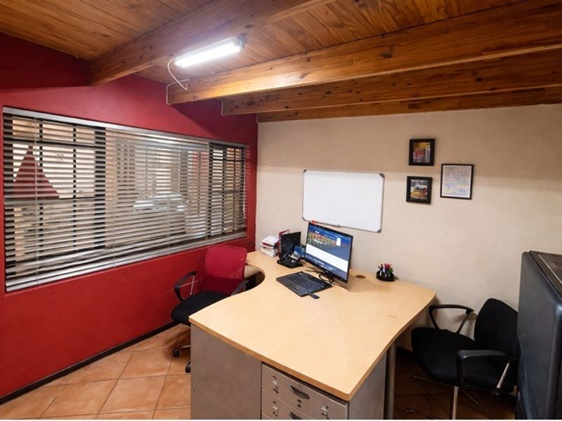 A functional home office featuring exposed timber ceiling beams, tiled flooring, and large windows with venetian blinds.