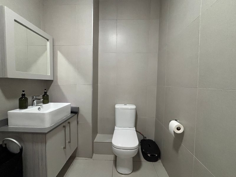 A modern bathroom featuring a vanity cabinet with a countertop basin, wall-mounted mirror, and a standard toilet, finished with neutral wall and floor tiles.