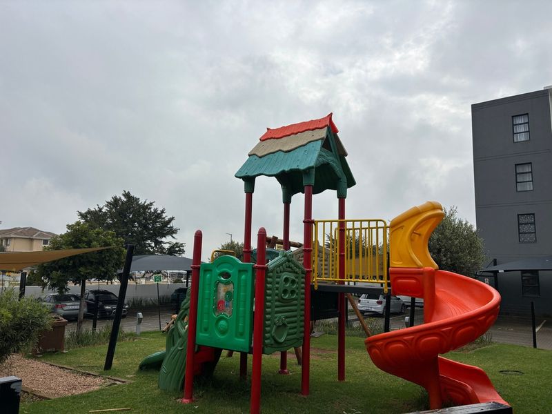 Communal garden area featuring a children's playground structure set within a landscaped lawn, adjacent to the apartment complex parking facilities.