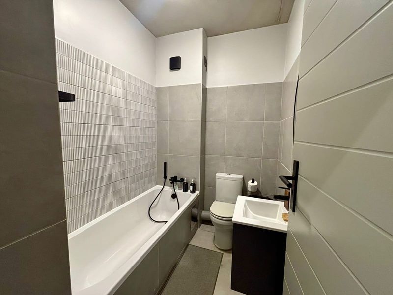 A modern, well-maintained bathroom featuring contemporary grey wall tiling, a bathtub with black matte fixtures, a vanity basin, and a standard toilet.