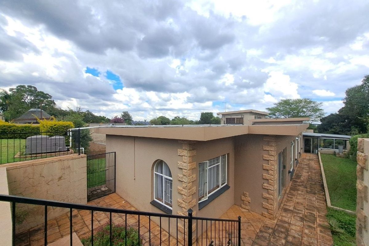 Rear view of the property featuring a tiled patio, stone-clad accents, flat roof with solar panels, and surrounding garden area with security fencing.