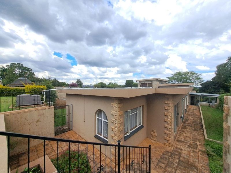 Rear view of the property featuring a tiled patio, stone-clad accents, flat roof with solar panels, and surrounding garden area with security fencing.