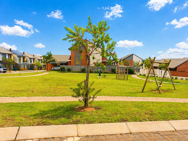 A well-maintained communal garden area within a townhouse complex, featuring paved walkways, manicured lawns, and established residential units in the background.