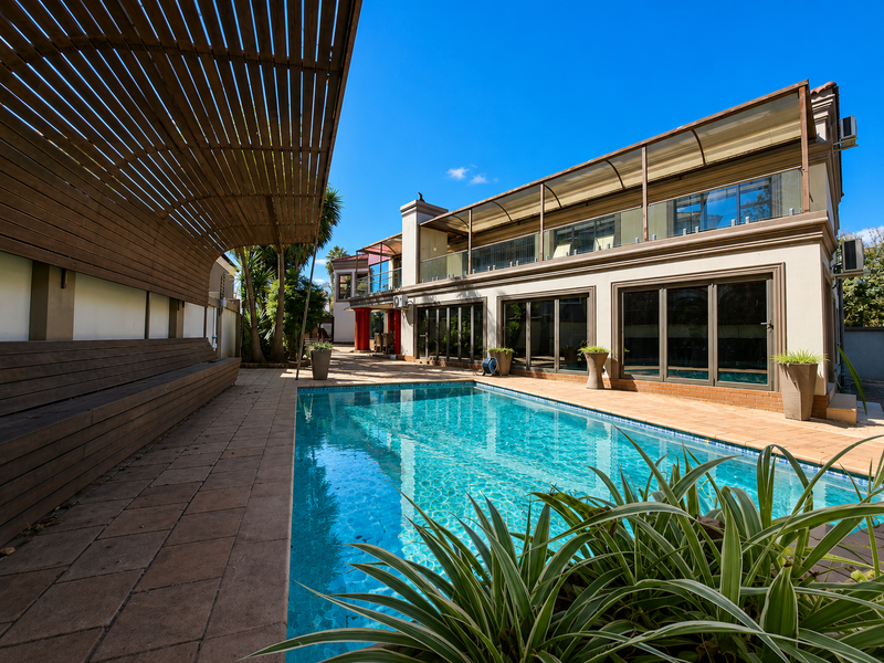 A well-maintained outdoor pool area featuring a modern swimming pool, a large wooden pergola with built-in seating, and paved patio space.