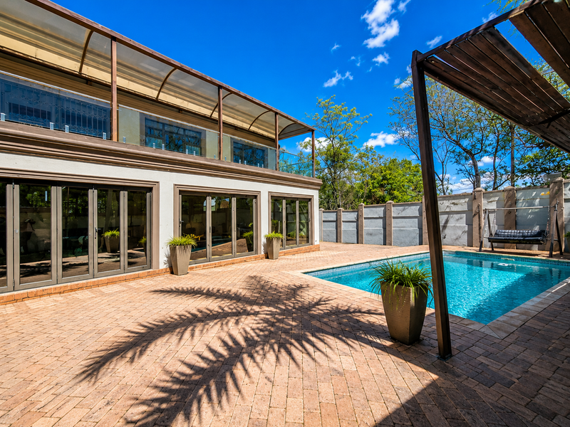 A well-maintained outdoor pool area featuring a paved patio, timber pergola, and large folding glass doors leading into the residence.