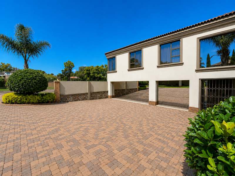 A well-maintained front exterior featuring a spacious paved driveway, brick boundary wall, and a modern building facade with large windows.