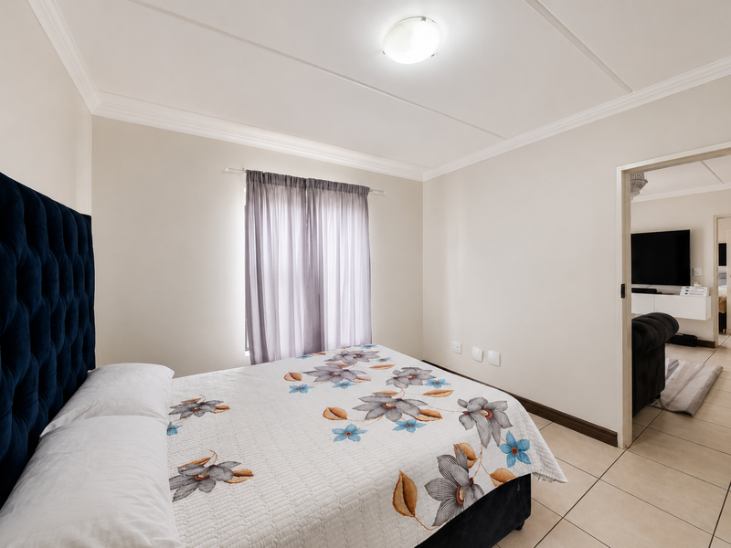 A well-maintained bedroom featuring neutral walls, tiled flooring, and a ceiling light, with a view into the adjacent living area.