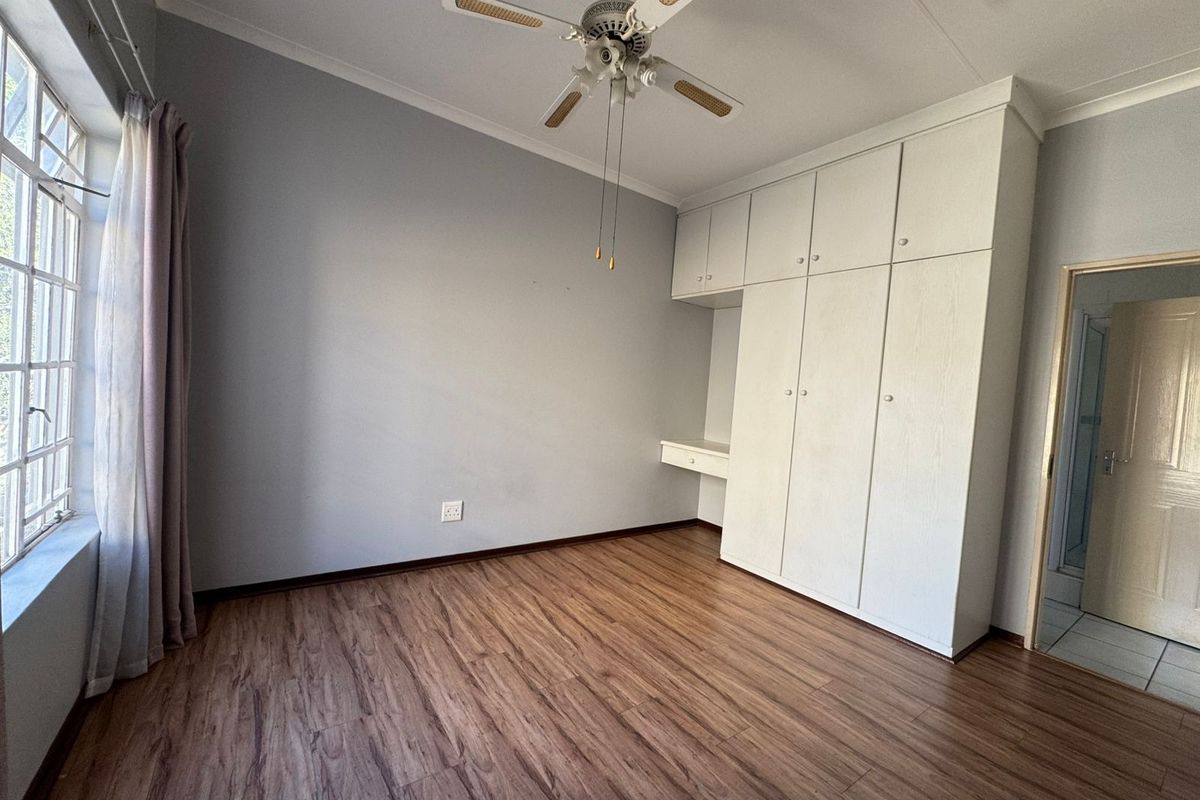 A bright, empty bedroom featuring laminate flooring, a ceiling fan, and ample built-in cupboard space with an integrated desk area.