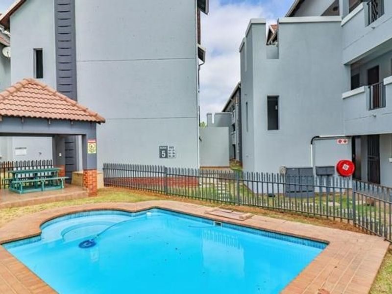 Communal swimming pool area featuring a paved surround, metal fencing, and a covered thatched gazebo for outdoor relaxation within the apartment complex.