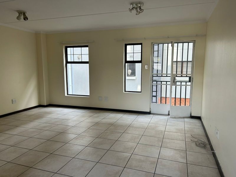 An empty, open-plan living area featuring neutral tiled flooring, ceiling-mounted lighting, and a secure metal entrance door leading to the exterior.