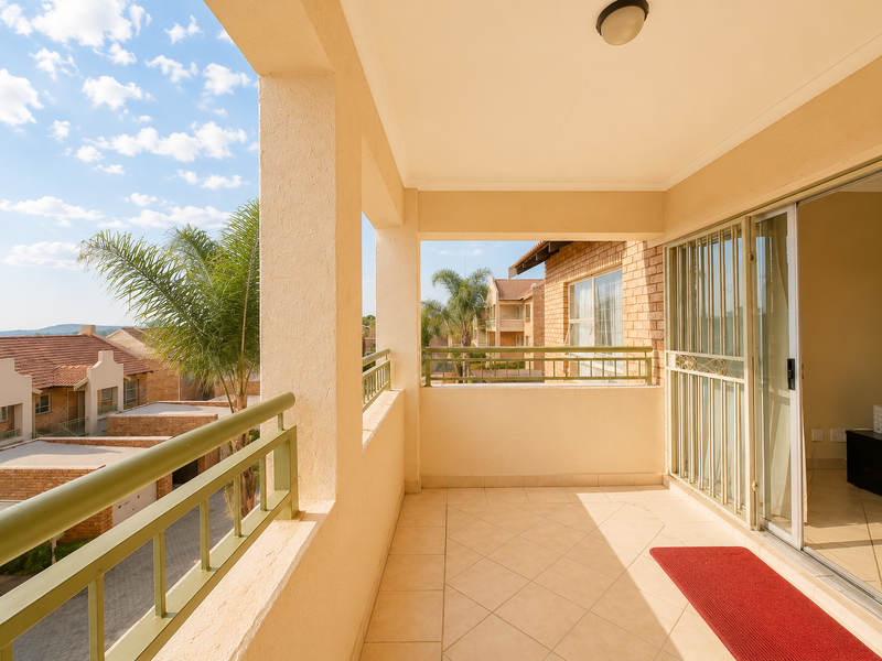 A covered balcony featuring tiled flooring, a metal balustrade, and a sliding glass door with a security gate, offering views of the surrounding townhouse complex.