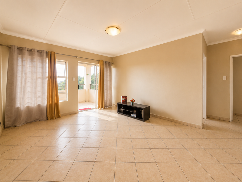 A spacious, sunlit living area featuring neutral tiled flooring, coved ceilings, and direct access to an outdoor balcony through a sliding glass door.