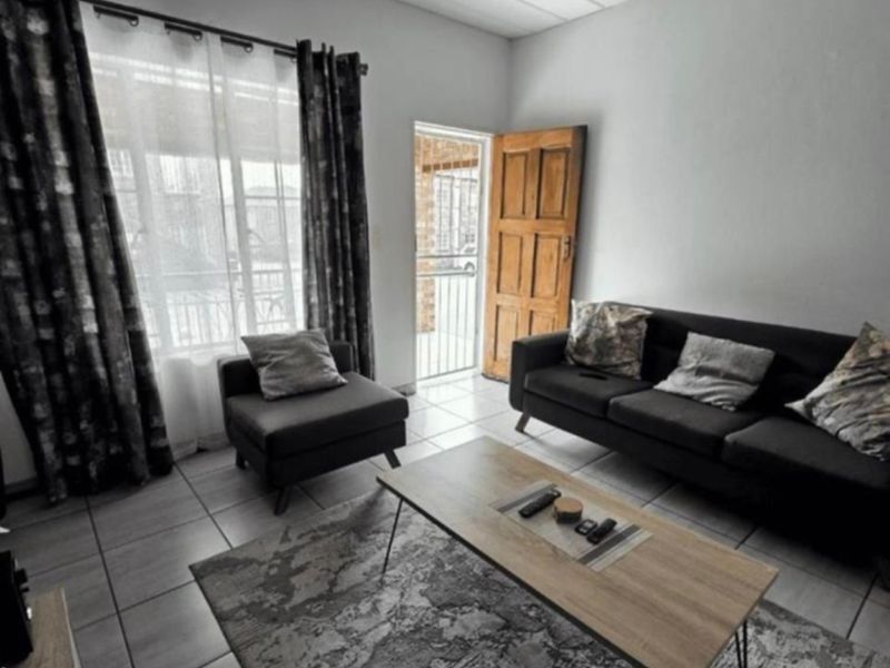 A functional living room featuring tiled flooring, a timber entrance door with security gate, and natural light through a large window.