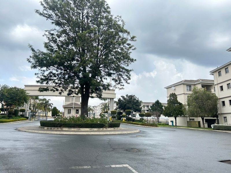View of the secure residential complex entrance featuring a landscaped roundabout, paved driveway, and gated security access point.