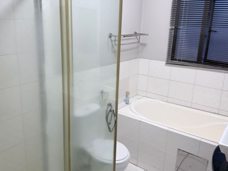 A functional bathroom featuring a built-in bathtub, separate shower enclosure, and toilet, finished with neutral wall tiling and a window with blinds.